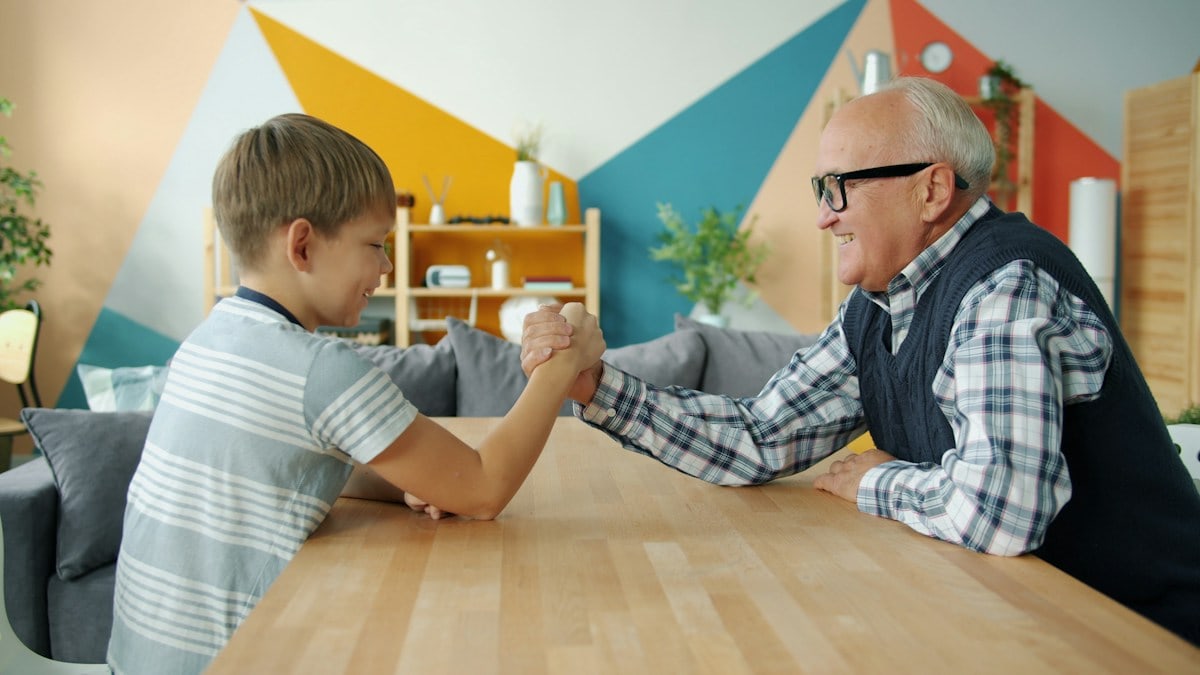 Caring grandfather is teaching boy to wrestle having fun doing sports at home, laughing grandad is winning talking to kid. Family, leisure time and lifestyle concept.
